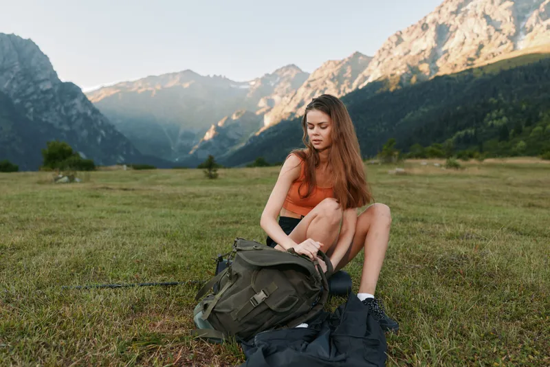 A young woman sits on grass beside a backpack in a scenic mountain valley. She wears an orange top and shorts, looking relaxed and thoughtful in the serene outdoor setting.