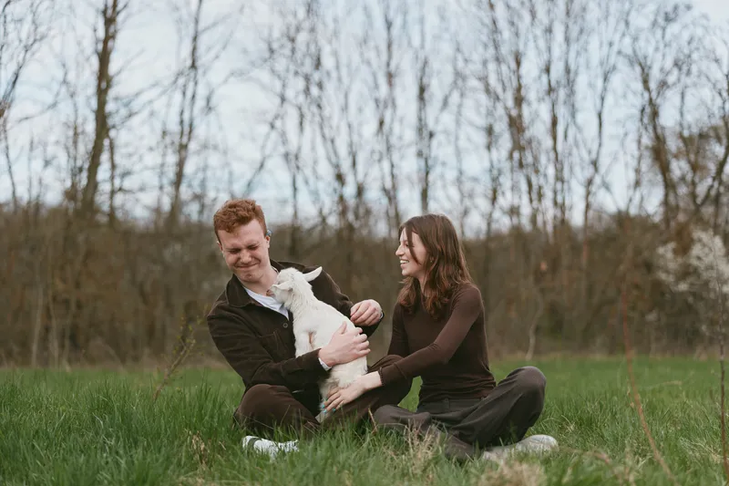 A joyful couple outdoors with a playful goat kid, sharing a light moment on a grassy field. A lifestyle scene highlighting bonds, nature, and gentle animal companionship, capturing warmth and