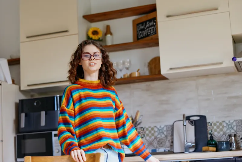 A woman with curly hair and glasses stands in a bright home kitchen, wearing a multicolor striped sweater. She leans on a counter, exuding casual confidence and everyday creativity.
