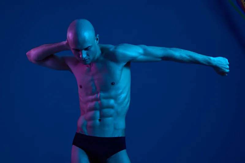 Athletic man posing in dramatic light, showcasing defined muscles. The image features a blend of blue tones, highlighting strength and vitality in a striking manner.