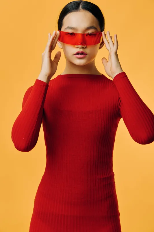 Woman wearing red dress and futuristic red sunglasses on yellow background. Portrait of stylish Asian female model posing with confident expression and trendy fashion look.