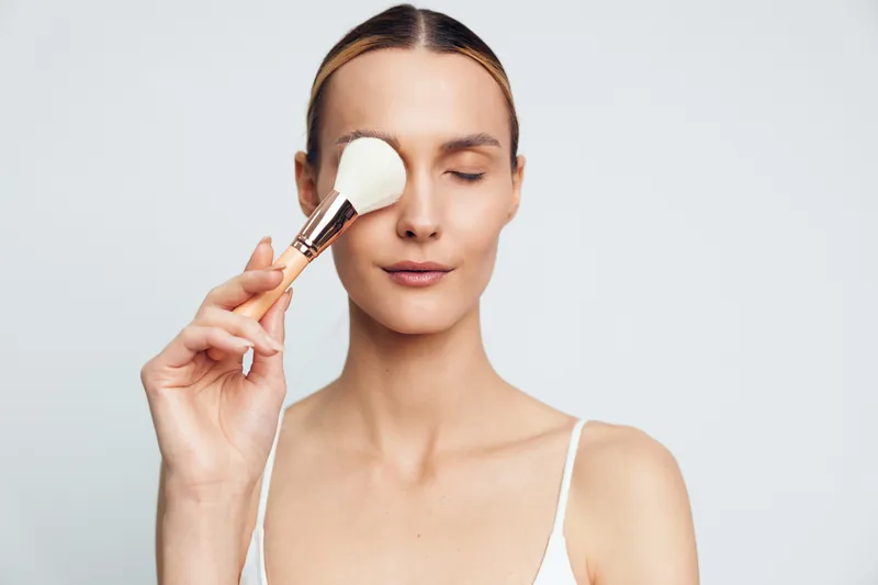 Young woman with clear skin applies powder makeup on face using brush with closed eyes isolated on white background, beauty routine and natural look concept.