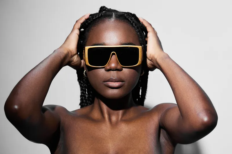 A confident Black woman with braided hair holding her head, wearing oversized yellow sunglasses, against a minimalist gray background Stylish and bold expression