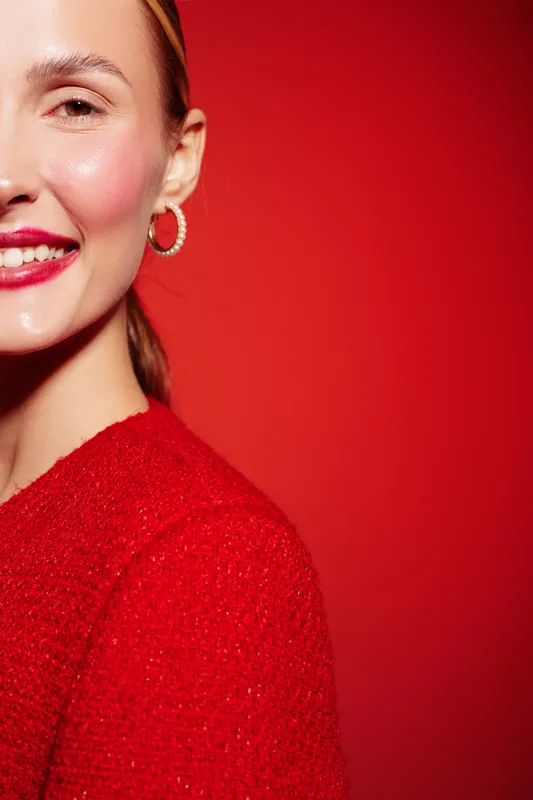 Close-up of smiling woman with red lipstick and hoop earrings wearing red sweater against red background. Concept of beauty, fashion, and confident female portrait with vibrant colors.