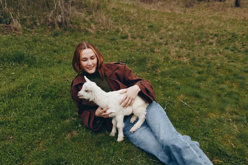 A relaxed woman lying on green grass gently cuddles a white goat kid, capturing a tender outdoor moment. A lifestyle scene showing connection with animals and simple countryside joy.