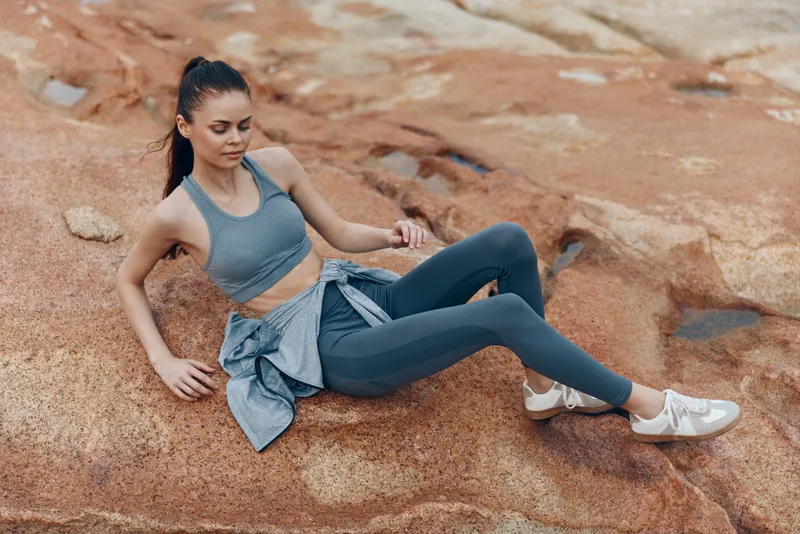 Young athletic woman in sportswear relaxing on rocky terrain, showcasing healthy lifestyle and fitness, expressing calmness in muted tones