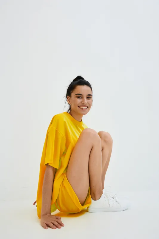 Smiling woman in yellow oversized t-shirt sitting on white floor with casual white sneakers, expressing happiness and relaxed mood in minimalistic bright studio setting.