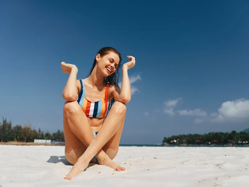 Young woman sits on white sand beach, legs crossed, arms raised in joy, wearing a bright striped swimsuit. Clear blue sky and calm sea frame a sunny moment.