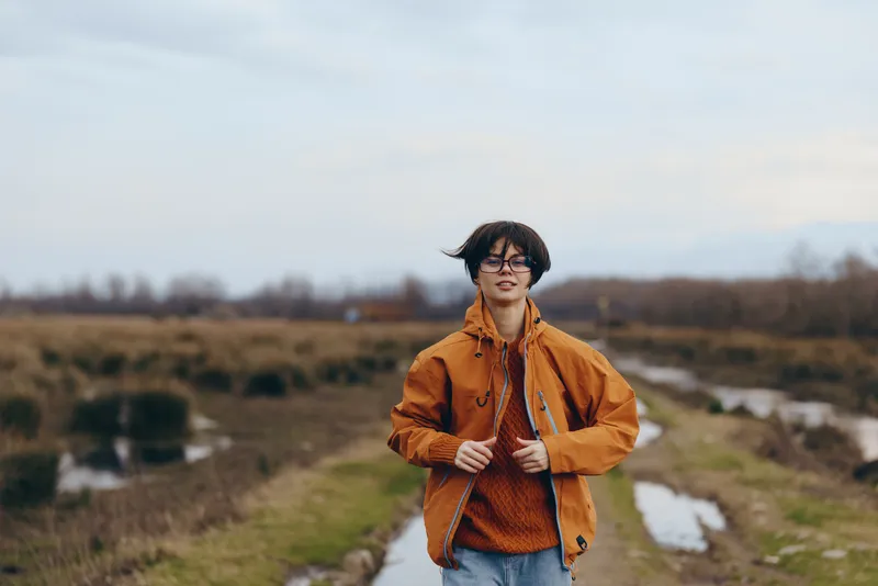 Woman outdoor lifestyle portrait, walking on a rural path in autumn, wearing orange jacket and glasses, relaxed posture, holding zipper at chest, countryside field and cloudy sky, soft blur background