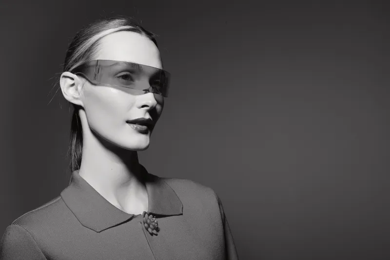 Portrait of a fair skinned Caucasian woman wearing a futuristic transparent visor, dressed in a vintage style coat against a dark background.