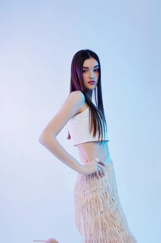Young woman with long hair wearing a white crop top and fringed skirt poses confidently against a light background. Concept of beauty, fashion, and style.
