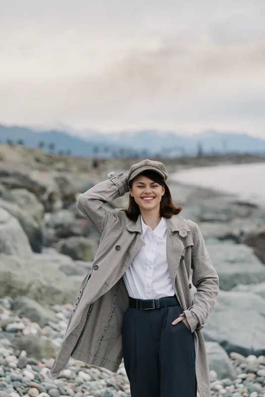 Smiling young woman in a grey coat and cap stands on a rocky beach with mountains in the background. Casual autumn fashion and outdoor lifestyle concept with natural scenery.