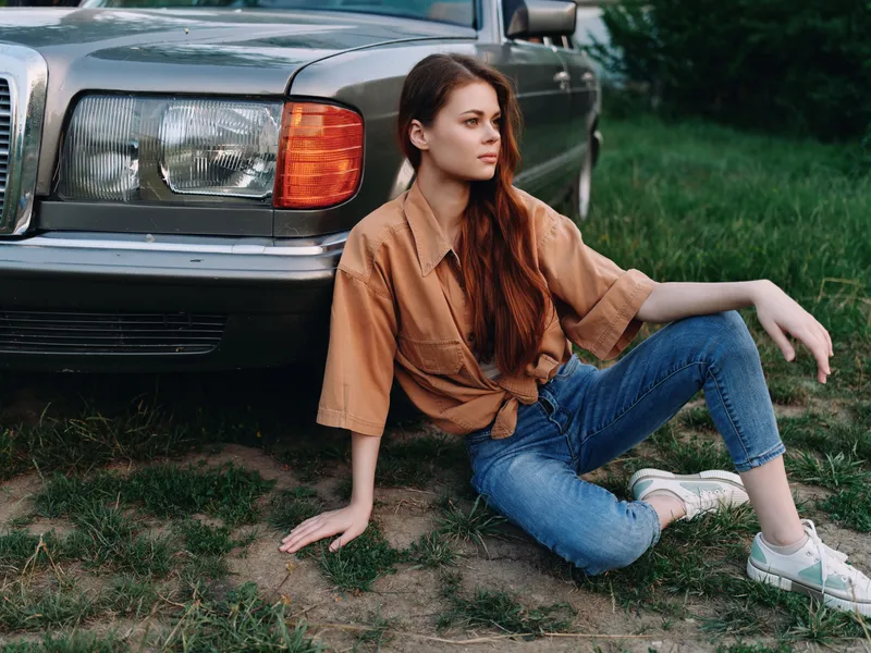 Young woman in casual fashion sitting beside a vintage car, expressing relaxed attitude in an outdoor setting with greenery, perfect for lifestyle concepts.