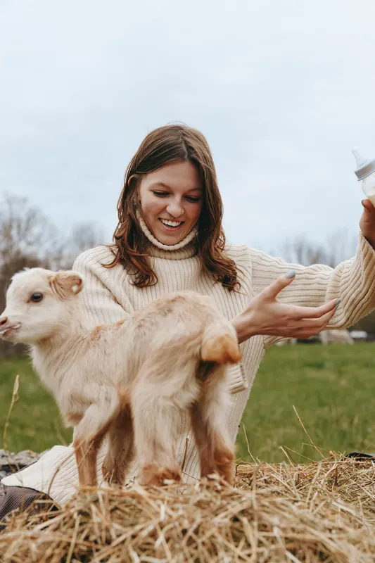 Young woman in a cozy sweater feeds a baby goat with a bottle outdoors on a cloudy day in a natural lifestyle setting with grass and hay around.