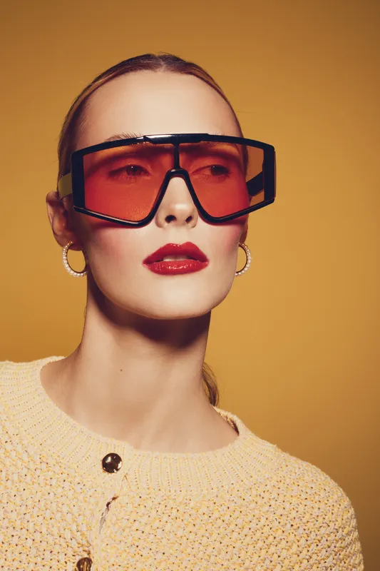 Stylish woman wearing oversized orange sunglasses and hoop earrings poses against a warm yellow background. Fashion portrait with glossy red lips and textured knitwear.