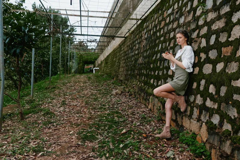 Young woman leaning against a stone wall in an outdoor setting, wearing a white blouse and a green skirt, exuding a relaxed and contemplative mood in a natural environment