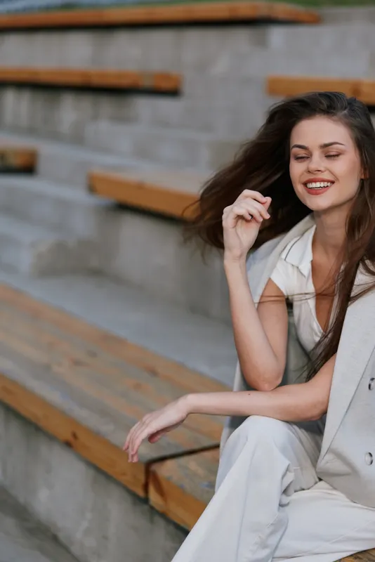 Confident woman in business attire smiling while sitting on wooden steps, representing empowerment and modern professionalism, showcasing a serene backdrop.