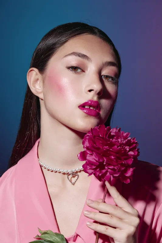 Young woman with bright pink makeup and glossy lipstick poses with a pink flower on a blue background. Fashion and beauty creative concept with vibrant colors.