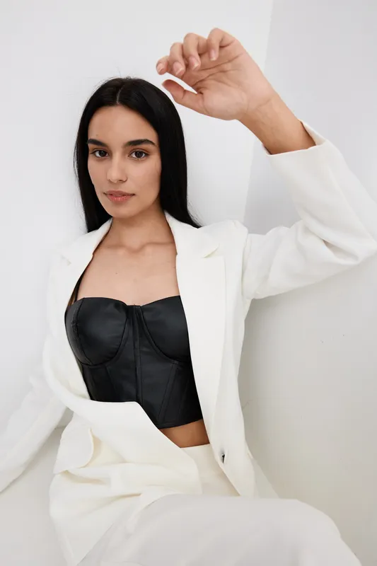young woman in white suit and black corset posing against white wall, natural look with straight dark hair, minimal makeup, indoor portrait with soft lighting