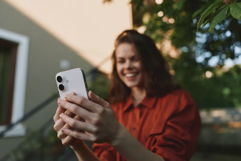 Smiling woman in casual orange shirt taking a selfie outdoors, framed by sunlight and greenery. Focus on lifestyle moments, self expression, and everyday connection