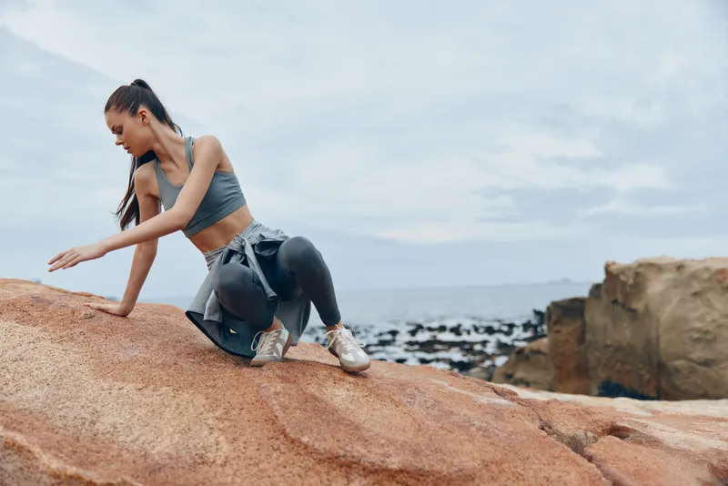Young athletic woman in stylish sportswear poses on rocky beach, showcasing a healthy lifestyle and fitness with a serene ocean background