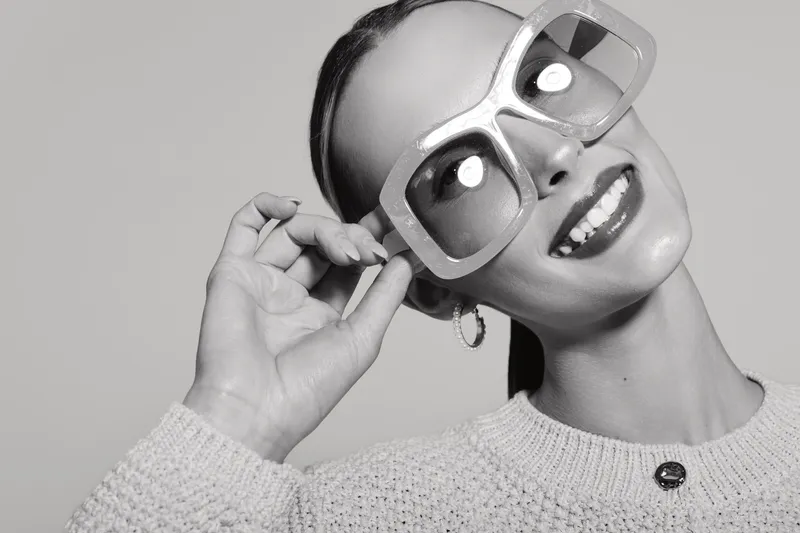Young Latin woman with medium skin tone smiles wearing oversized sunglasses and knit sweater. Studio portrait in black and white with joyful mood and fashion concept.