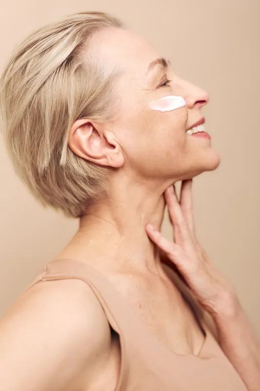 Smiling mature woman with short blonde hair applies white face cream on cheek, touching neck with hand. Concept of skincare, beauty, self-care, aging, wellness, and natural glow.