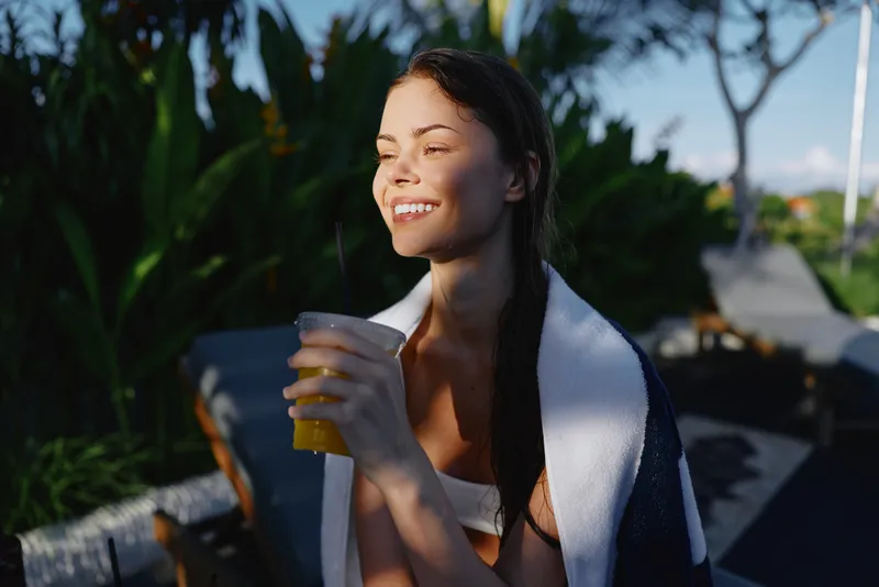Cheerful woman outdoors, holding a cold drink, smiling as sunlight warms her face, towel draped over shoulders, relaxed pose, lush garden and tropical backdrop, bright colors and gentle breeze