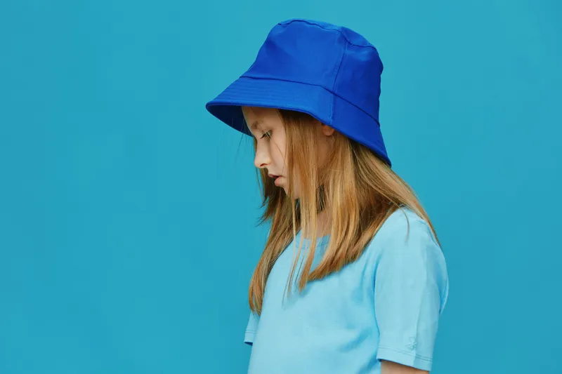 Profile of a young girl wearing a blue bucket hat and a matching t shirt against a turquoise background, conveying a calm, thoughtful emotion