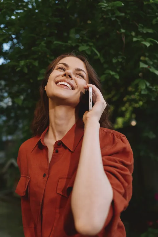 Smiling woman outdoors talks on phone in casual red shirt, showcasing a modern lifestyle moment with natural background and relaxed posture, bright daylight and energy