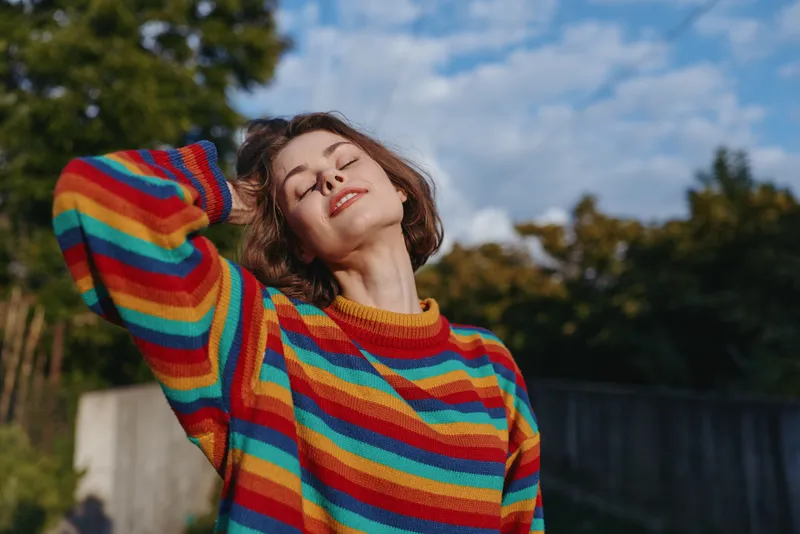 Young woman in her 20s wearing a rainbow sweater, short hair, smiling with eyes closed outdoors, leaning back in sunlight in a casual outfit lifestyle portrait conveying relaxation and joy.