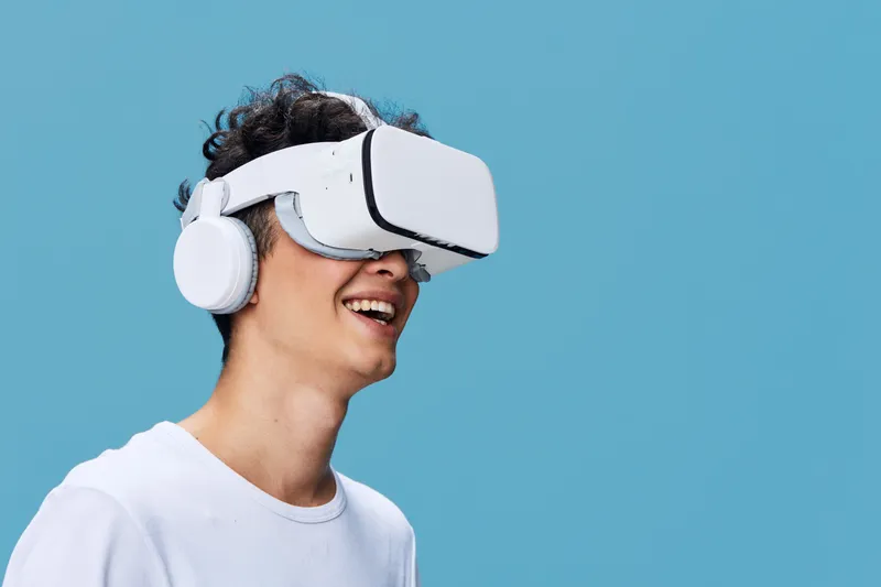 young man wearing white virtual reality headset and headphones, smiling with excitement, isolated on blue background, immersive technology experience