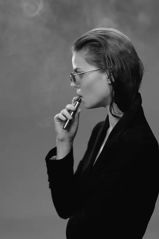 Black and white profile of a stylish woman vaping with sleek hair and sunglasses in a smoky atmosphere.