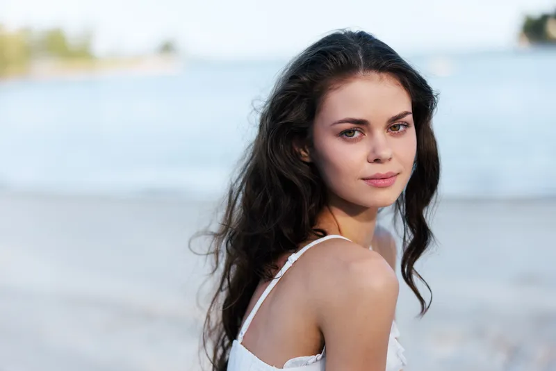 Portrait of a young woman with dark hair on a sandy beach, wearing a light top, gazing softly at the camera as gentle coastal light frames her calm expression.