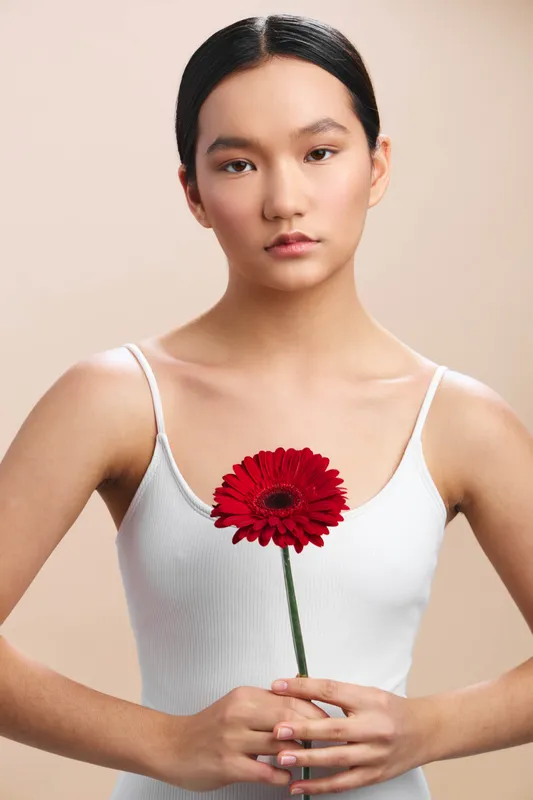 asian woman stands gracefully in a minimalist studio, holding a single red flower, calm and poised, conveying quiet elegance and focus in a simple portrait.