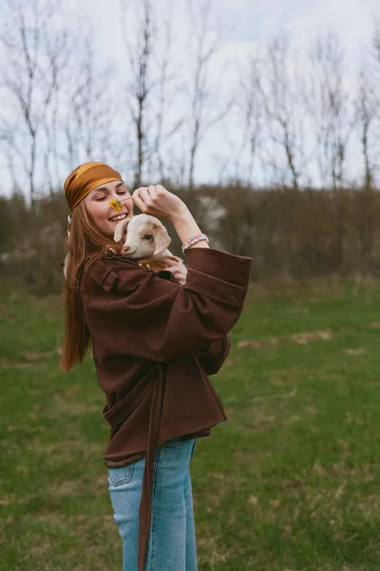 Cheerful woman outdoors, holding a playful goat kid, lifestyle moment in natural light, affection and smiling joy, warm breeze, denim and brown coat, meadow backdrop, rustic charm, candid connection