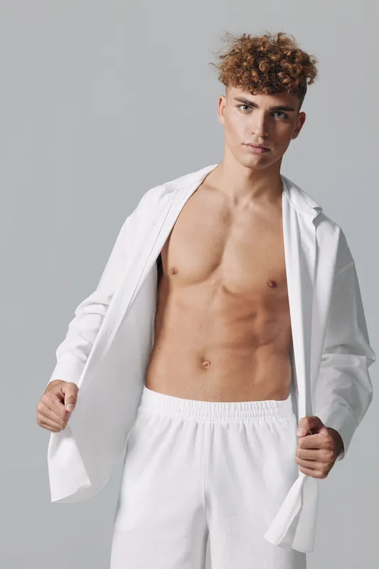 Young man with curly hair posing shirtless wearing white open shirt and white pants against gray background. Fashion model with toned body and confident expression in studio.