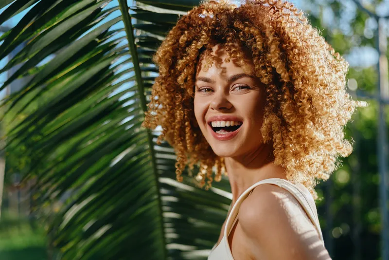 smiling woman with curly hair enjoying sunny outdoor environment near tropical palm leaves, natural light, youthful and cheerful expression