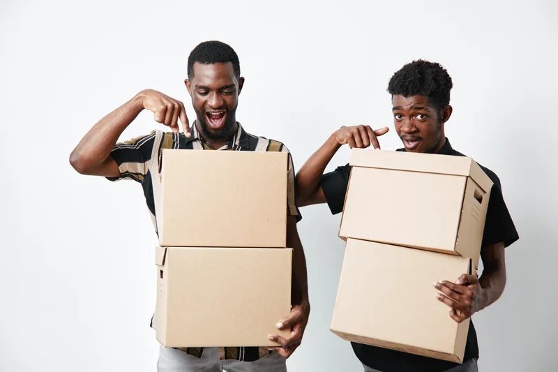Two african american men with dark skin holding cardboard boxes for delivery and moving, smiling and pointing at packages on white background in studio.