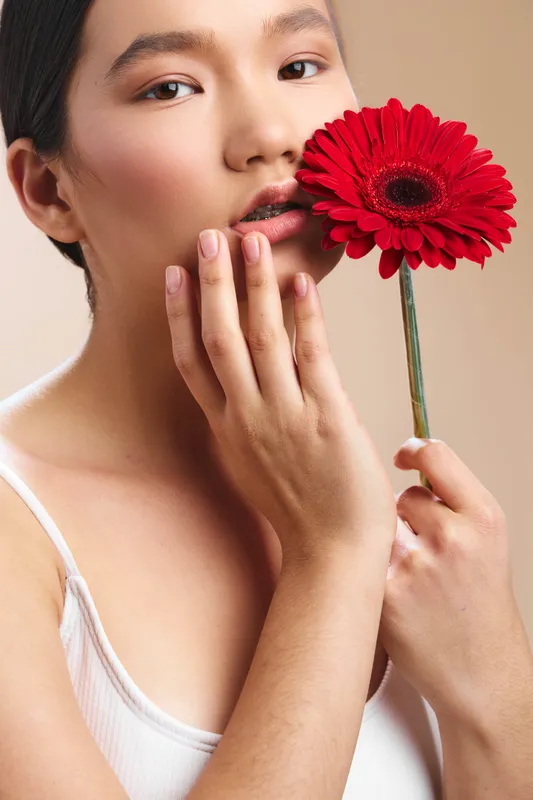 asian woman gently holds a vibrant red flower near her face, soft lighting highlights delicate features, calm expression, and refined pose creating an elegant studio portrait for beauty and lifestyle