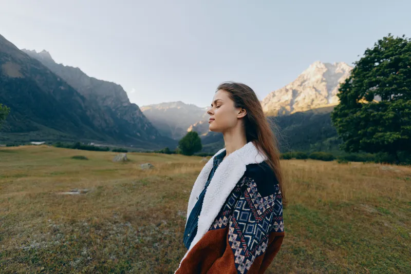 Woman standing in meadow facing mountains, nature portrait outdoors at sunrise. Cozy patterned sweater suggests autumn travel and peaceful solitude with fresh air and scenic valley view.