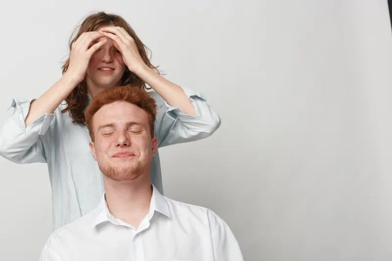 Young man with closed eyes enjoys head massage from woman standing behind him. Relaxation and stress relief concept in casual clothing on neutral background with soft natural light.