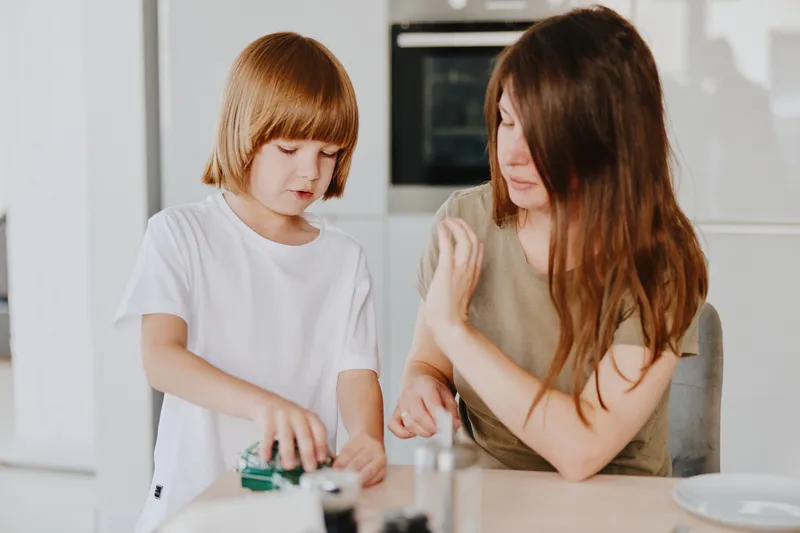 Caring woman engaged in a fun activity with a child, promoting creativity and bonding in a bright, modern kitchen setting.