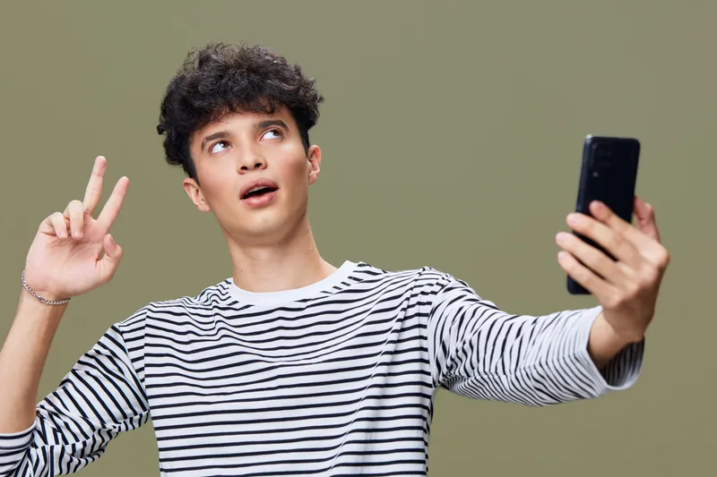 young man taking selfie with smartphone making peace sign wearing striped shirt on neutral background with curious expression