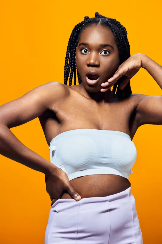 Shocked young woman with braided hair in a strapless top and pastel pants, posing against a vibrant yellow background, expressing surprise and excitement
