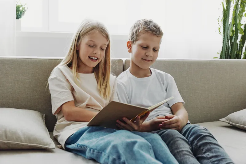Two children sitting on sofa reading a book together, enjoying learning and sharing a quiet moment in bright natural light living room with casual clothing and cozy atmosphere.
