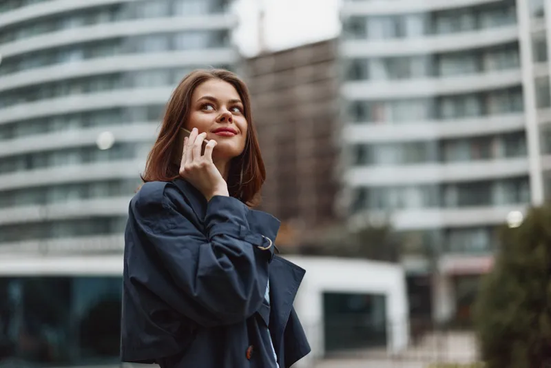 A thoughtful woman in a coat stands outdoors in an urban cityscape, talking on a phone, with modern buildings behind, capturing casual communication in daily life.