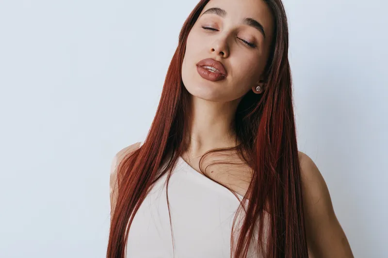 Young woman with long red hair and closed eyes posing in white top against light background. Natural beauty, calm expression, and relaxed mood in studio portrait with soft lighting.