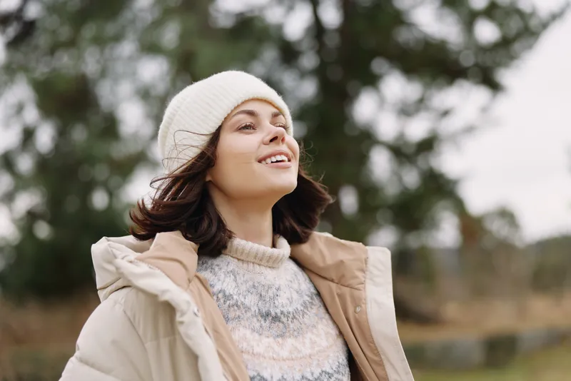 Woman outdoors, smiling in a knit beanie and warm coat in a breezy winter landscape, close up portrait highlighting comfort, resilience and natural light in a peaceful setting.
