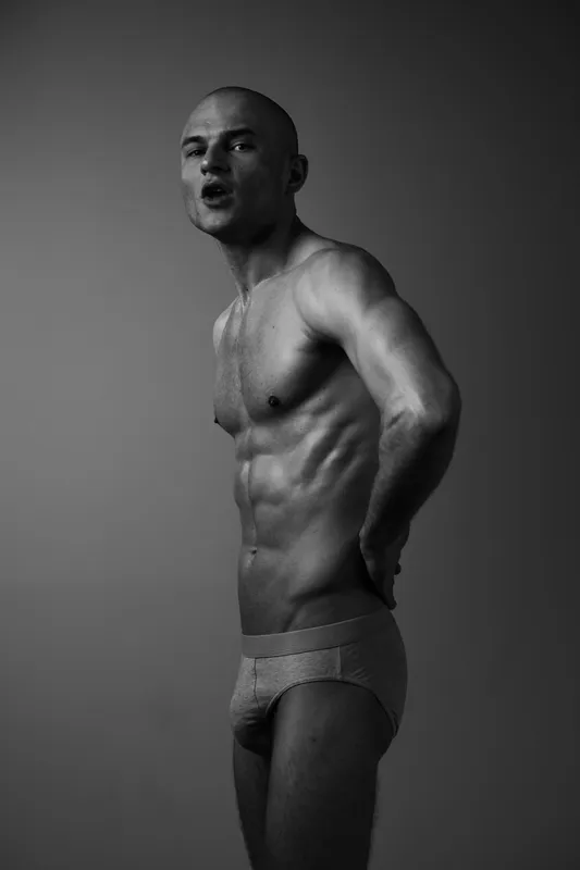 Black and white image of a man showcasing strength and confidence, emphasizing muscular physique and expressive pose against a neutral backdrop.